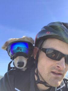 Photo of Eric wearing a bike helmet with his dog on his bike in a bike helmet and goggles