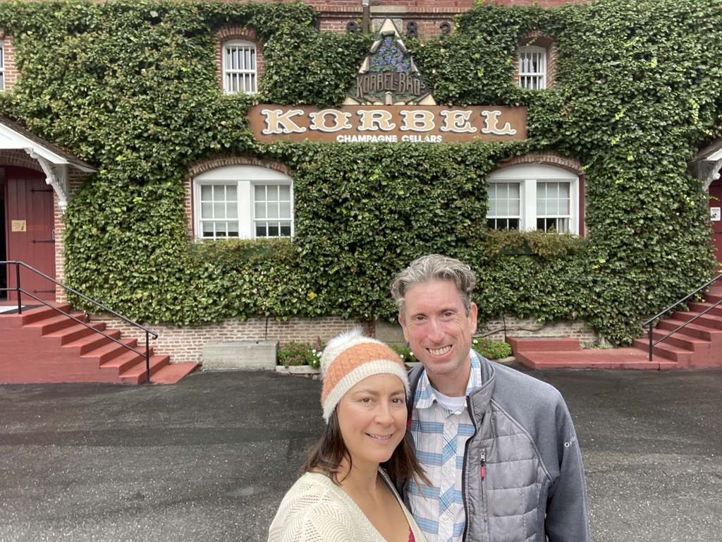 Jessica and her husband stand in front of an ivy-laden building with "Korbel" on the sign. Jessica is wearing a striped beanie hat and a white shirt, and her husband is wearing a plaid button-down shirt with a gray jean jacket on top.