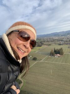 Jessica leans out of a hot air balloon, high above rolling fields of green and trees, wearing sunglasses and a beanie hat