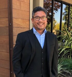 Man wearing blue shirt and black suit jacket in front of brown building and vegetation, wearing glasses